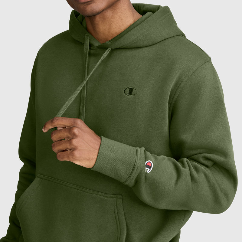 A person wearing an olive green Champion Powerblend Hoodie, C Logo adjusts the drawstring. The Champion logo is visible on the chest and sleeve of the men’s fleece hooded sweatshirt. The person’s face is partially out of frame.
