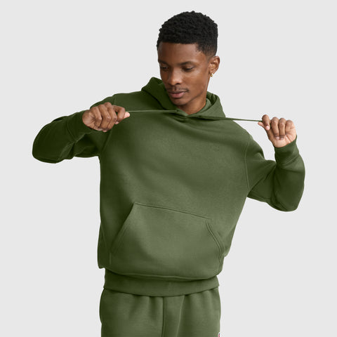 A person wearing a green Champion Powerblend Hoodie, C Logo, and matching sweatpants pulls on the hooded sweatshirt drawstrings while looking down. The background is plain light gray.