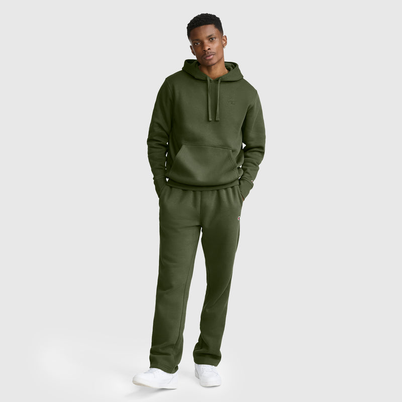 A person stands against a plain background wearing a Champion Powerblend Hoodie, C Logo in olive green with matching sweatpants and white sneakers, hands in pockets, looking at the camera.