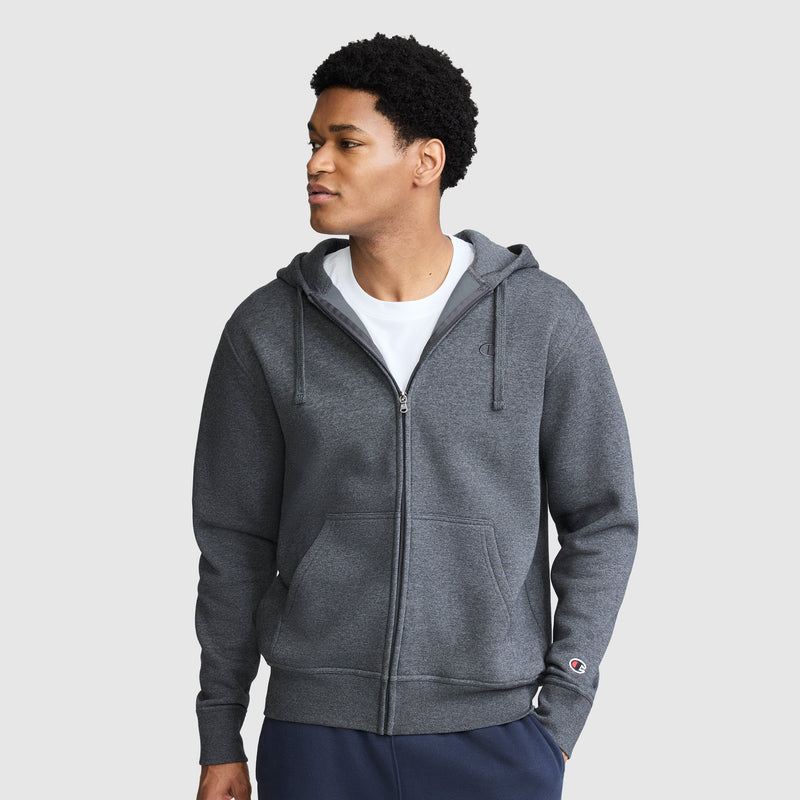 A man wearing a Champion Powerblend Full-Zip Hoodie, C Logo in dark gray over a white T-shirt stands against a plain light gray background, looking to his left with his hands in his pockets, dressed comfortably in Champion men's sweatpants.