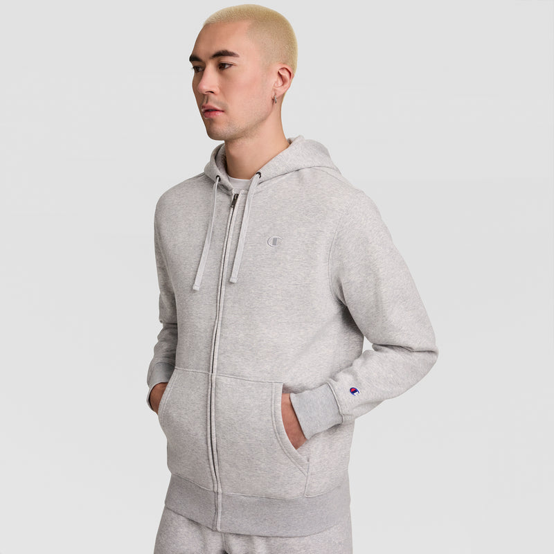 A person with short blond hair wears a light gray Champion Powerblend Full-Zip Hoodie, featuring the C Logo, and matching pants, standing against a plain white background with hands in pockets.