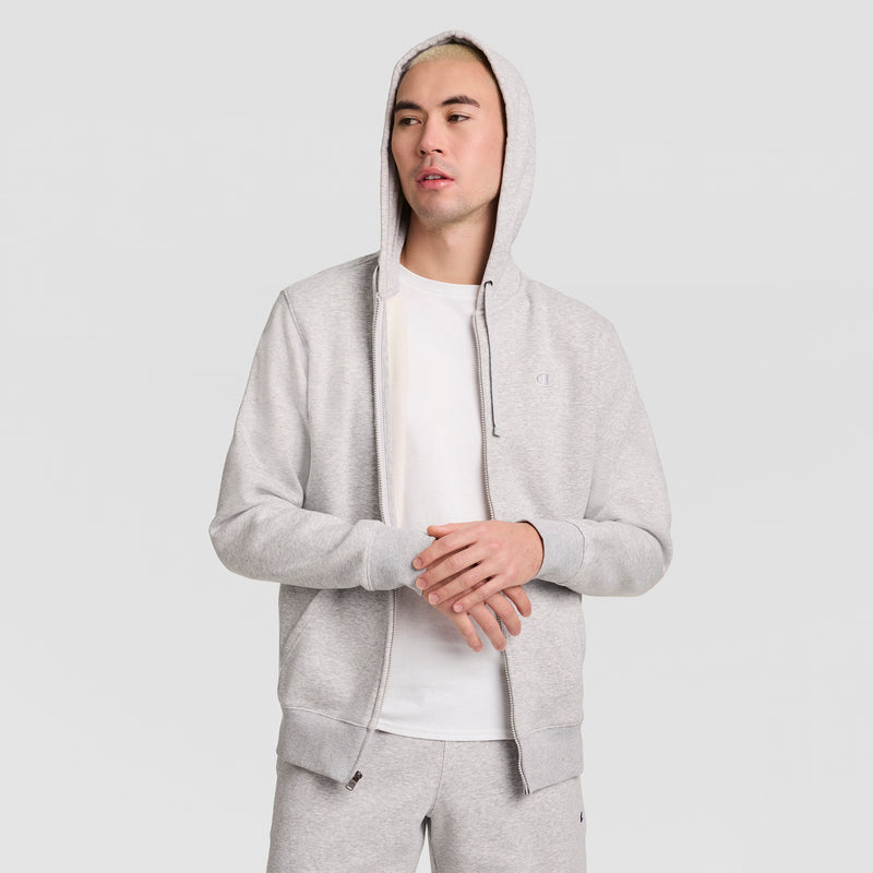 A person wearing a light gray Champion Powerblend Full-Zip Hoodie, C Logo, stands against a plain light background, gazing slightly to the side with hands loosely clasped.