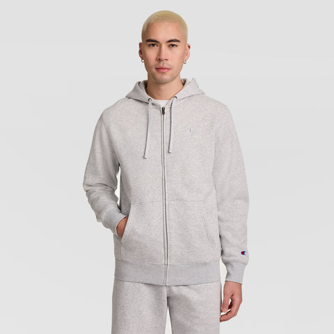 A person with short blond hair stands against a plain white background, wearing a light gray Champion Powerblend Full-Zip Hoodie, C Logo, paired with matching sweatpants and resting one hand in a pocket.