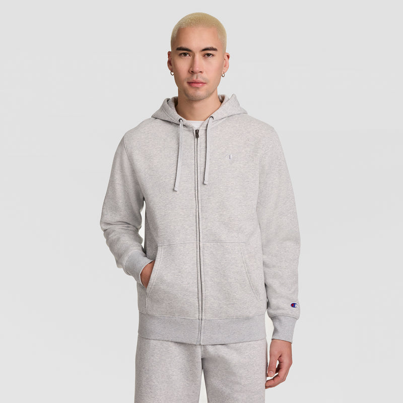 A person with short blond hair stands against a plain white background, wearing a light gray Champion Powerblend Full-Zip Hoodie, C Logo, paired with matching sweatpants and resting one hand in a pocket.