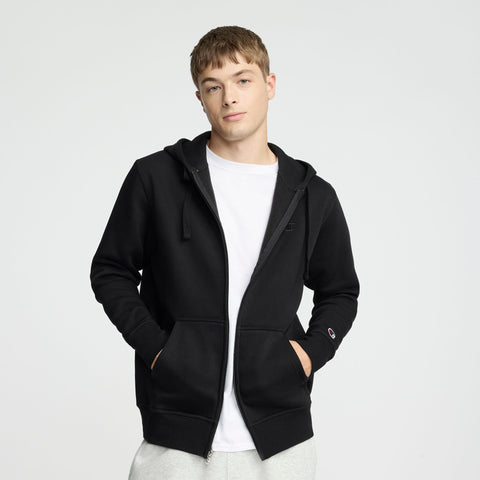 A young man wearing a Champion Powerblend Full-Zip Hoodie, C Logo, over a white t-shirt and light gray pants stands with his hands in his pockets against a plain light background.