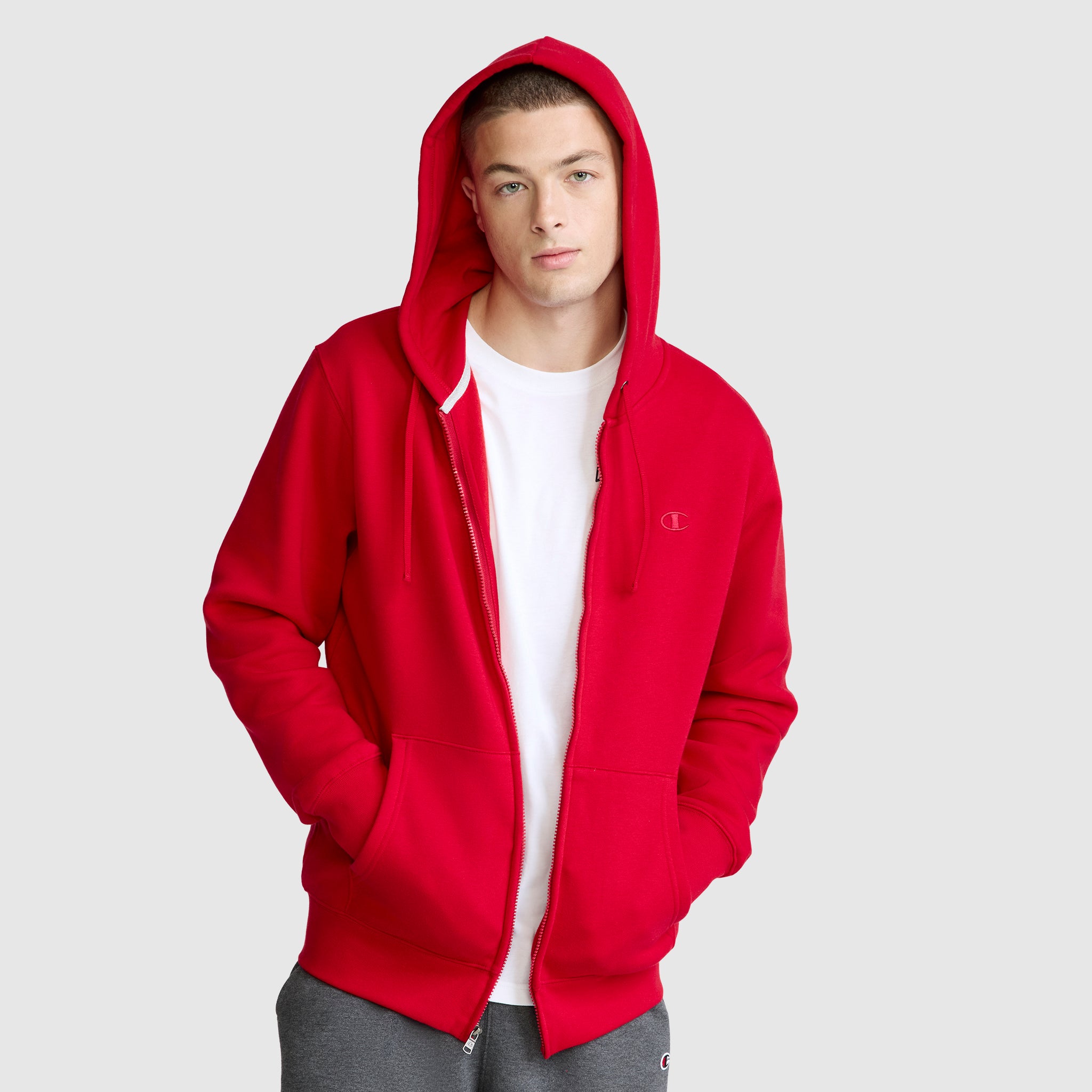 A young man stands with his hands in his pockets and the hood up, looking at the camera against a plain light gray background. He is wearing a Champion Powerblend Full-Zip Hoodie, C Logo in bright red over a white t-shirt, paired with Champion Men's Powerblend Sweatpants whose cinched bottoms add a modern touch to these comfortable fleece sweatpants.