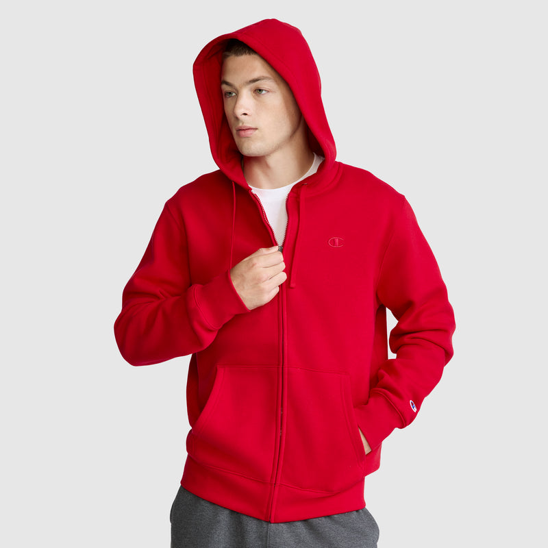 A person wearing a Champion Powerblend Full-Zip Hoodie, C Logo in bright red with the hood up and gray Champion Men's Powerblend Sweatpants stands against a plain light gray background. The hoodie features a small Champion "C" logo on the chest and a front pocket.