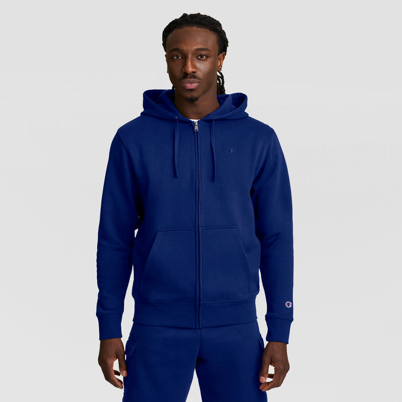 A man stands facing forward in a matching royal blue Champion Powerblend Full-Zip Hoodie, C Logo, and sweatpants. The hoodie features a hood, front pockets, and small embroidered logos on the chest and sleeve. The background is plain light gray.