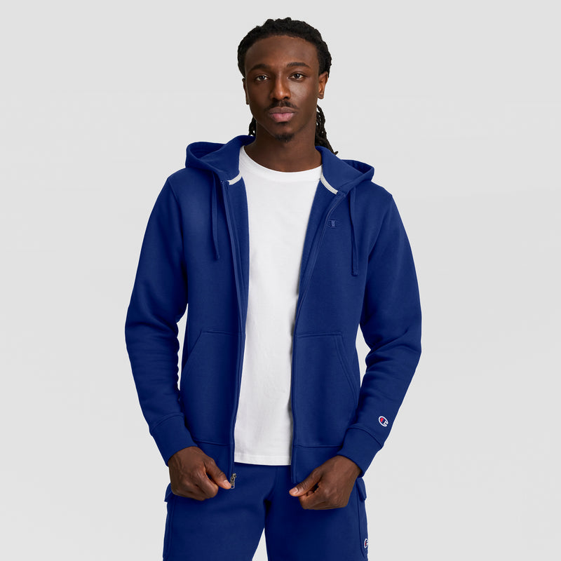 A man wears a Champion Powerblend Full-Zip Hoodie, C Logo, over a white tee and matching blue pants, standing forward with hands in pockets against a plain light gray background.