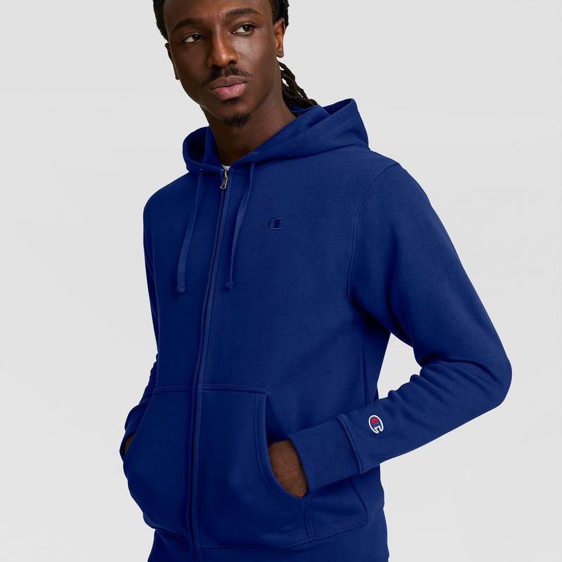 A man wearing a blue Champion Powerblend Full-Zip Hoodie, C Logo, with hands in pockets, looks slightly to the side. The sweatshirt has small embroidered logos on the chest and sleeve, set against a plain light gray background.