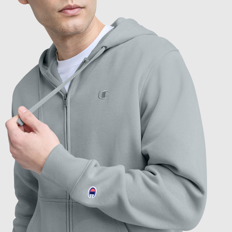 Someone wears a light gray Champion Powerblend Full-Zip Hoodie, C Logo, featuring a small C logo on the chest and a patch logo on the sleeve, while holding one of the drawstrings.