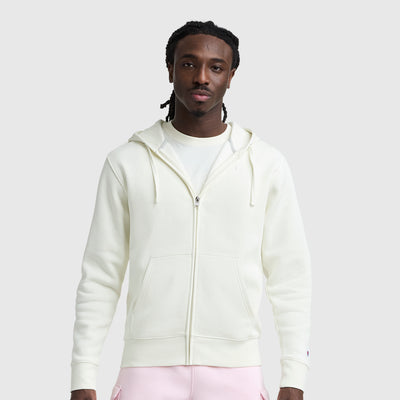 A man with braided hair faces the camera against a light gray background, wearing a cream Champion Powerblend Full-Zip Hoodie, C Logo, over a white t-shirt and light pink pants.