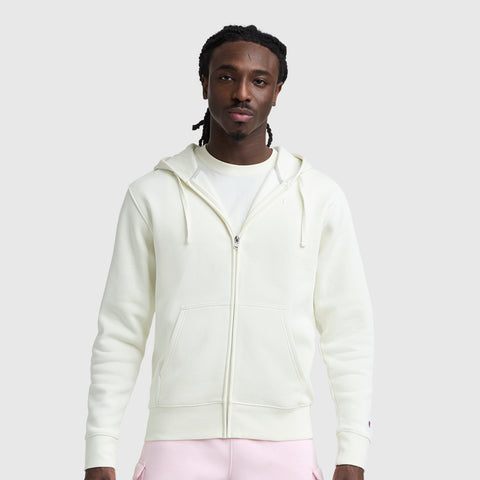 A man with braided hair faces the camera against a light gray background, wearing a cream Champion Powerblend Full-Zip Hoodie, C Logo, over a white t-shirt and light pink pants.