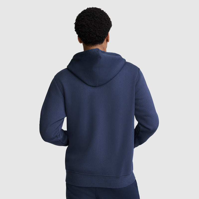 A person stands facing away from the camera, wearing a Champion Powerblend Full-Zip Hoodie, C Logo in dark blue with matching pants, against a plain light gray background.