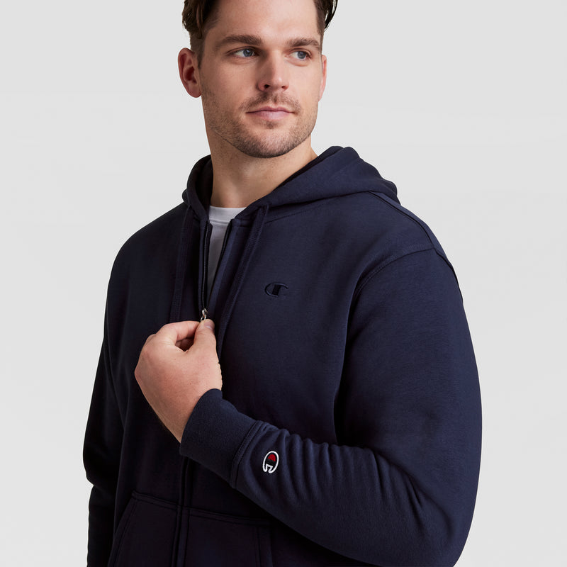 A man wearing a navy blue Champion Powerblend Full-Zip Hoodie, C Logo - Big & Tall, gazes to the side with one hand near the zipper. The hoodie features small Champion logo patches on the chest and left wrist. Plain light gray background.