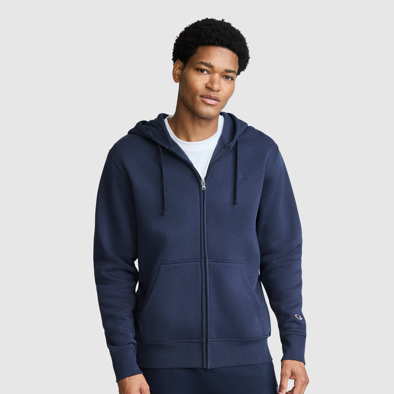 A man is shown facing forward against a plain light gray background, wearing a Champion Powerblend Full-Zip Hoodie, C Logo in navy blue with a hood and drawstrings, paired with matching pants.
