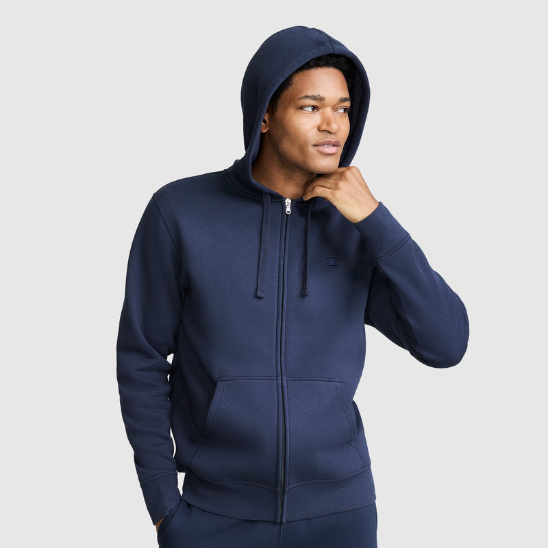A person wearing a Champion Powerblend Full-Zip Hoodie with the C logo in navy blue stands with one hand touching their chin, looking to the side against a plain light background.