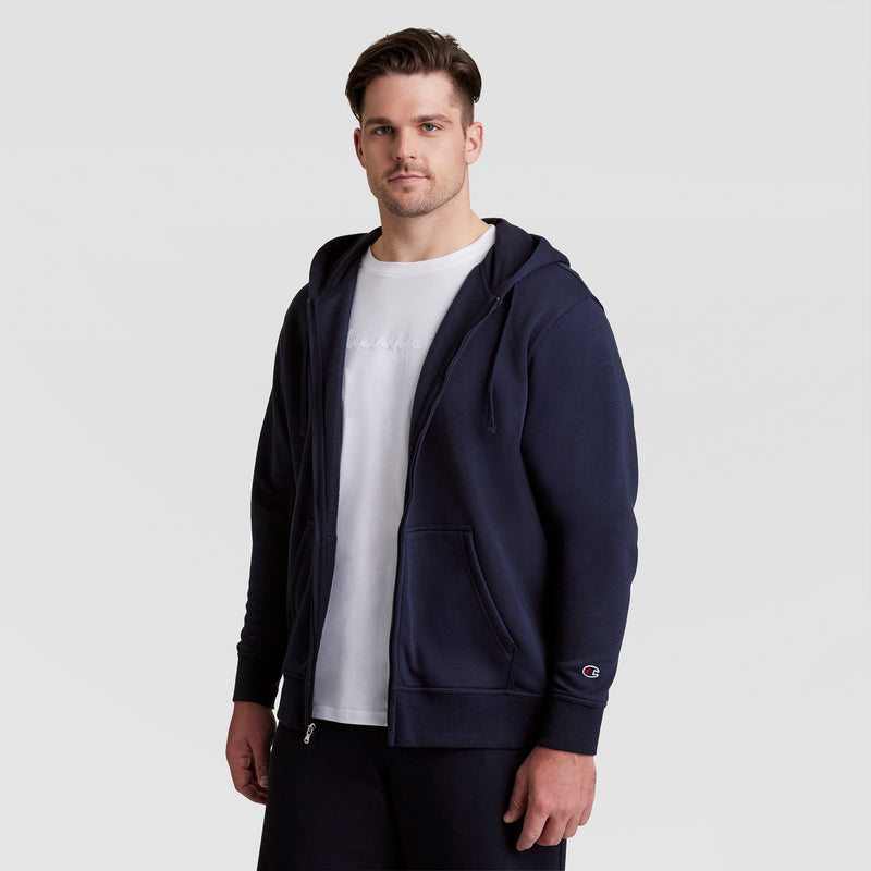 A man stands facing forward, wearing a navy Champion Powerblend Full-Zip Hoodie, C Logo - Big & Tall over a white t-shirt and black pants, against a plain light gray background.