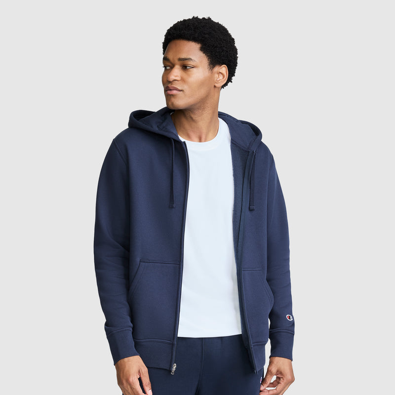 A man wearing a navy blue Champion Powerblend Full-Zip Hoodie, C Logo, styled over a white t-shirt, stands against a plain light gray background looking slightly to his left.