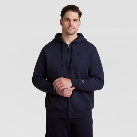 A man wearing the Champion Powerblend Full-Zip Hoodie, C Logo - Big & Tall in navy blue, featuring a hood and drawstrings, stands against a plain light gray background, looking at the camera with his hands clasped.