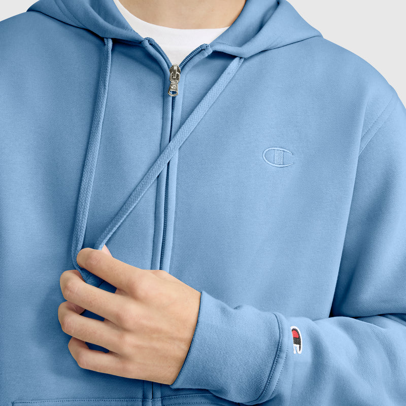 A person wears a light blue Champion Powerblend Full-Zip Hoodie, C Logo, holding one of the hoodie’s drawstrings. The person’s face is not visible.