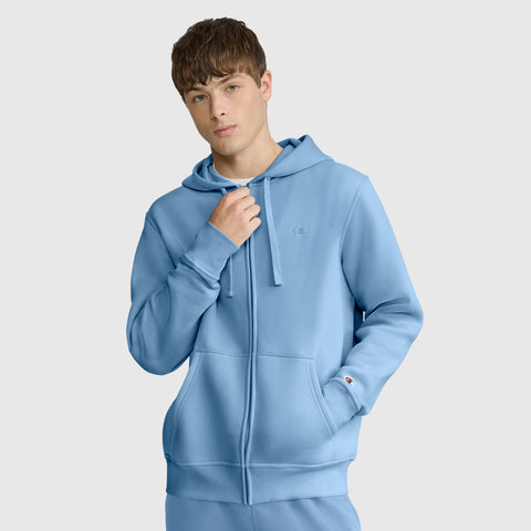 A young man wearing a light blue Champion Powerblend Full-Zip Hoodie with the C Logo stands against a plain light gray background, looking at the camera with a neutral expression.