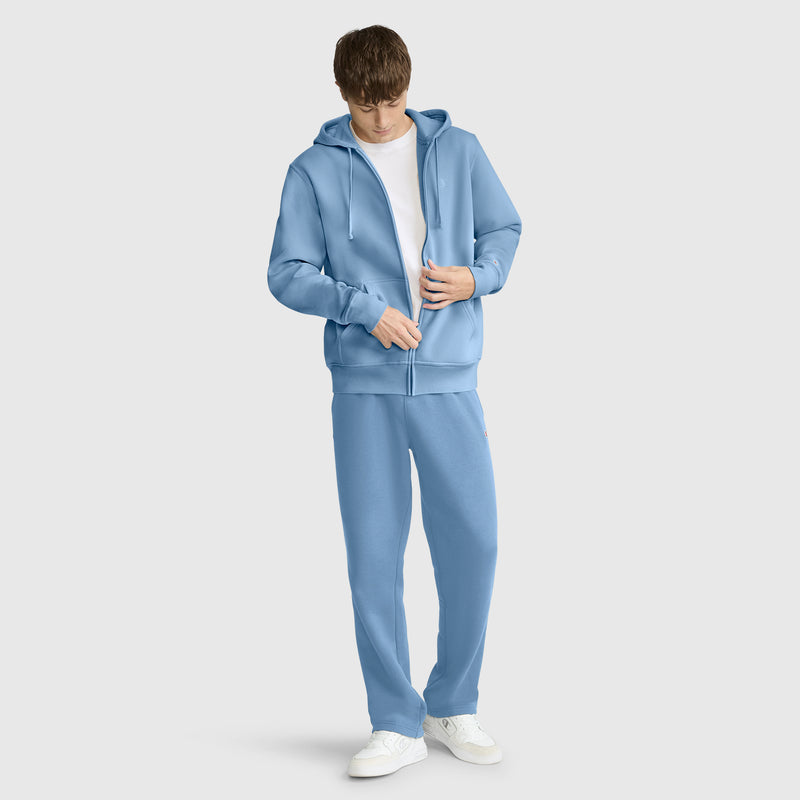 Wearing a light blue Champion Powerblend Full-Zip Hoodie with the signature C logo, paired with matching sweatpants over a white t-shirt, a person stands against a plain background looking down while adjusting the zipper on the hoodie.
