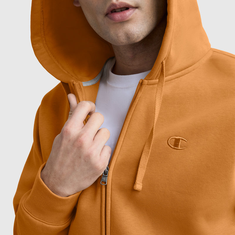 A person wears a Champion Powerblend Full-Zip Hoodie, C Logo in orange with the hood up and the zipper slightly open to reveal a white shirt underneath. The hoodie features an embroidered C logo on the chest.