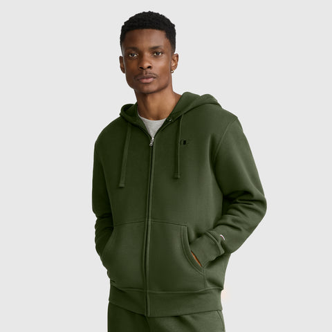 A man wearing a green Champion Powerblend Full-Zip Hoodie, C Logo—with a hood and drawstrings—stands with his left hand in his pocket, looking at the camera against a plain light gray background.