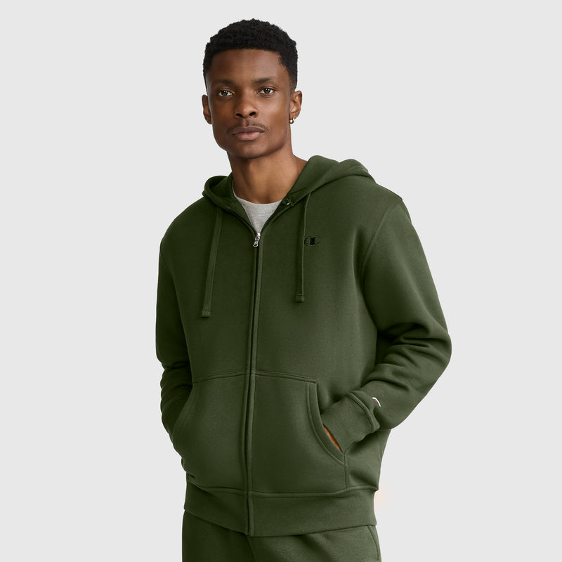 A man wearing a green Champion Powerblend Full-Zip Hoodie, C Logo—with a hood and drawstrings—stands with his left hand in his pocket, looking at the camera against a plain light gray background.