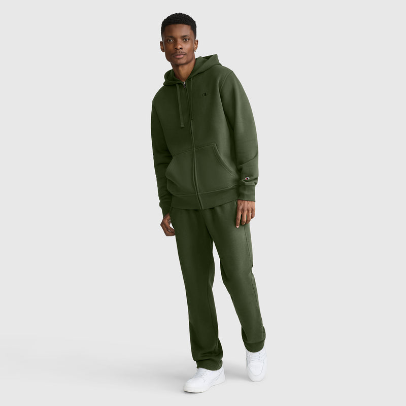A person stands against a plain light gray background wearing a dark green Champion Powerblend Full-Zip Hoodie with the C logo, paired with matching sweatpants and white sneakers.