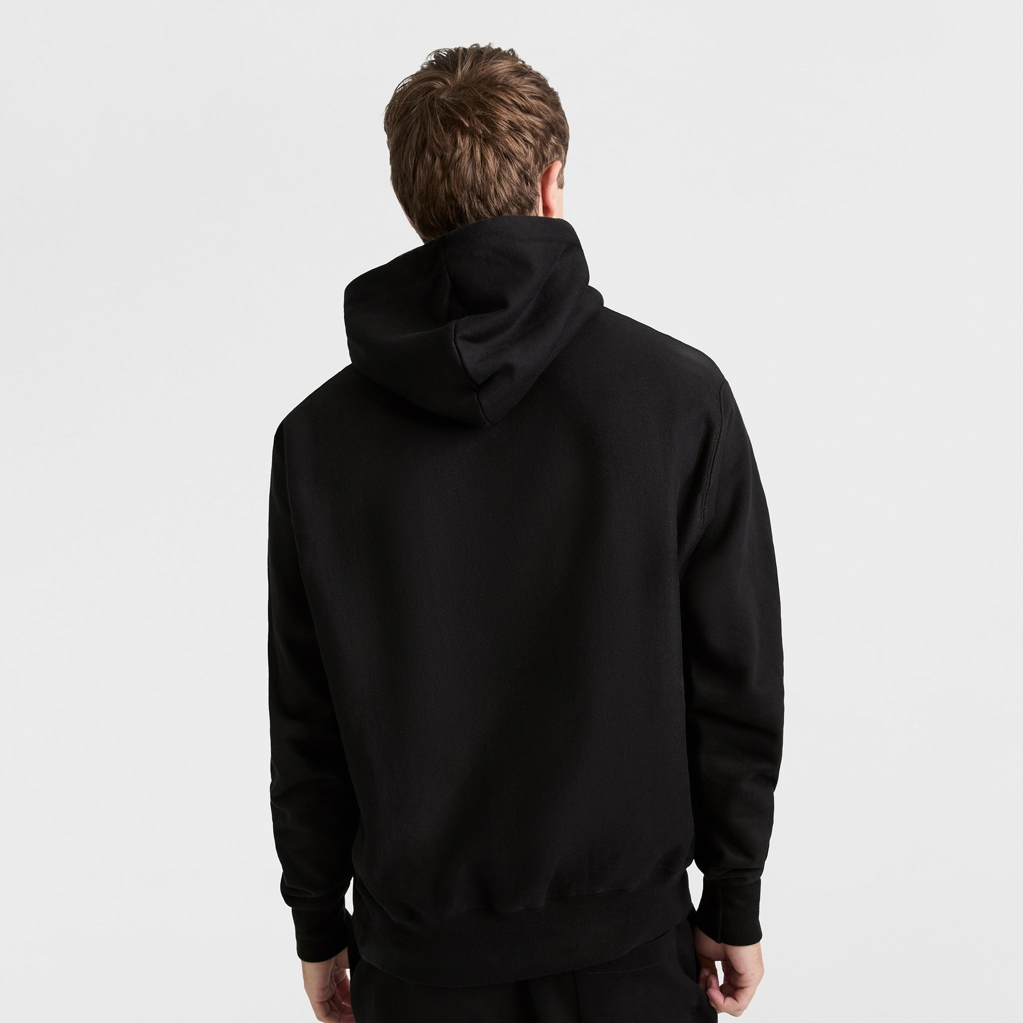 A person with short brown hair faces away from the camera, wearing a plain black Champion Reverse Weave Hoodie with the C Logo and the hood down, against a plain white background.