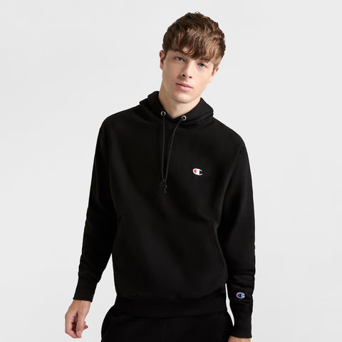 A young man with short brown hair wears a black Champion Reverse Weave Hoodie, C Logo—a heavyweight pullover with small chest and sleeve logos—standing against a plain light background.