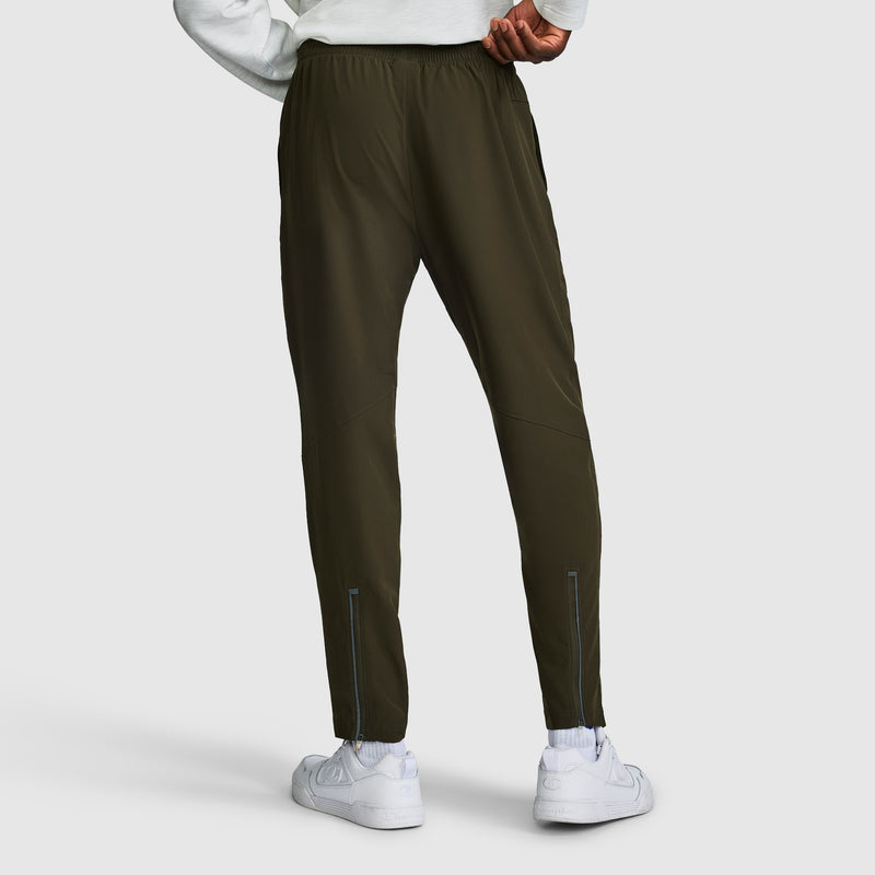 A person is shown from the back wearing dark green Champion MVP Pants, Moisture Wicking, C Logo men’s slim-fit sweatpants, paired with a white sweatshirt and white sneakers. The pants feature reflective details near the ankles and are crafted from moisture-wicking fabric for comfort during any activity.