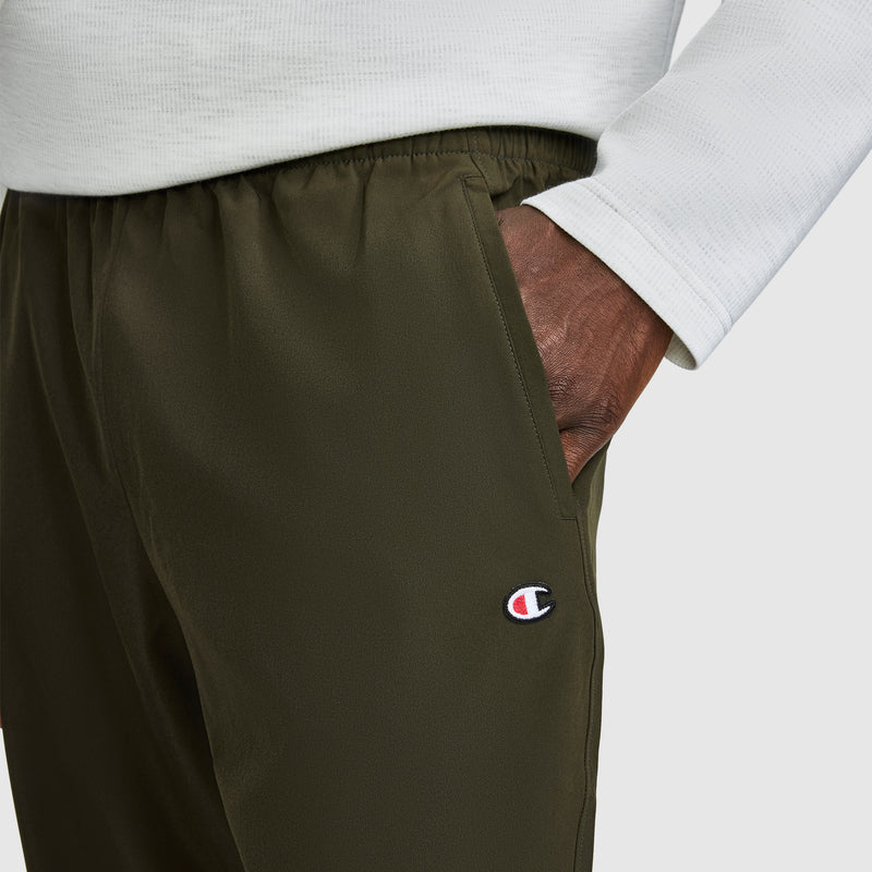 A person wearing Champion MVP Pants, featuring moisture wicking fabric and a C Logo in olive green, along with a white long-sleeve shirt, has their hand in their pocket.