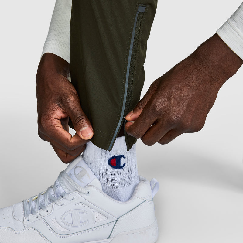 A person unzipping the ankle zippers of olive green Champion MVP Pants with moisture wicking and C logo features, revealing white Champion socks and white Champion sneakers on a light background.