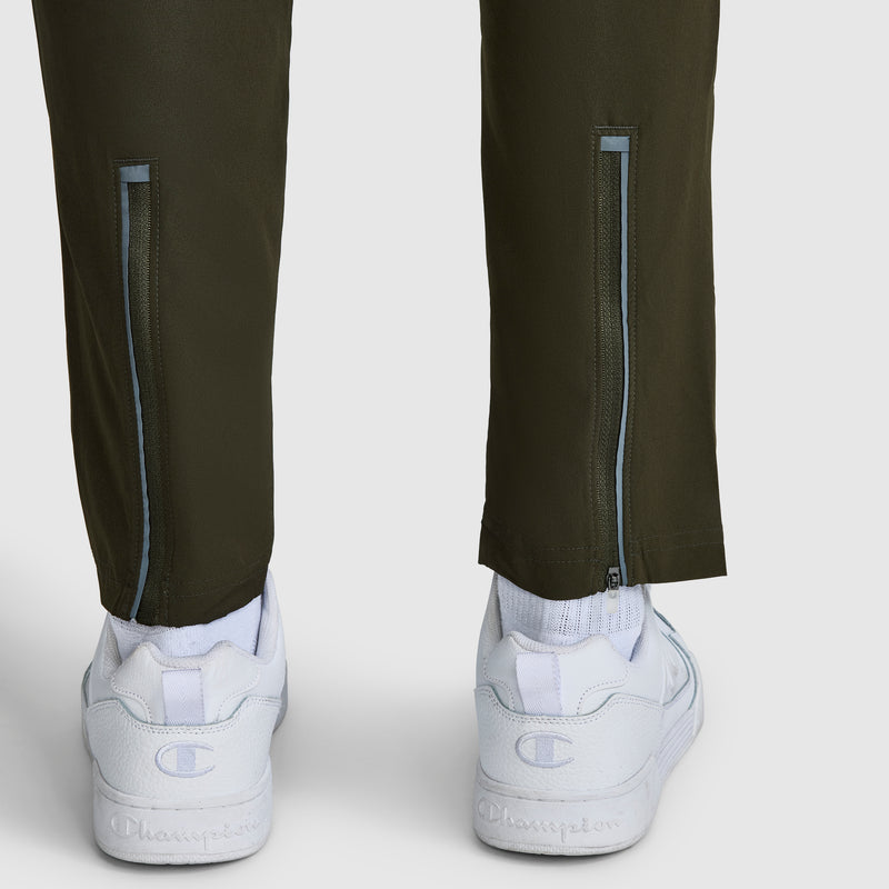 A close-up of the back of a person’s lower legs wearing Champion MVP Pants featuring a slim fit, moisture-wicking fabric, ankle zippers, and the signature C logo, paired with white Champion sneakers.