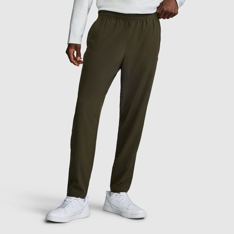 A person stands against a plain white background, showcasing the lower half of their outfit: olive green Champion MVP Pants with moisture-wicking fabric and ankle zippers. The men’s slim-fit joggers, featuring the signature C logo, are styled with a white long-sleeve shirt and white sneakers.
