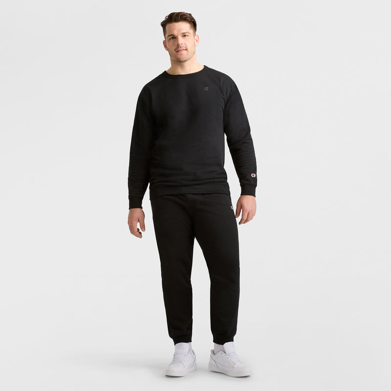 A man faces forward wearing Champion Powerblend Joggers, C Logo – Big & Tall in black with a black sweatshirt and white sneakers, set against a plain white background.