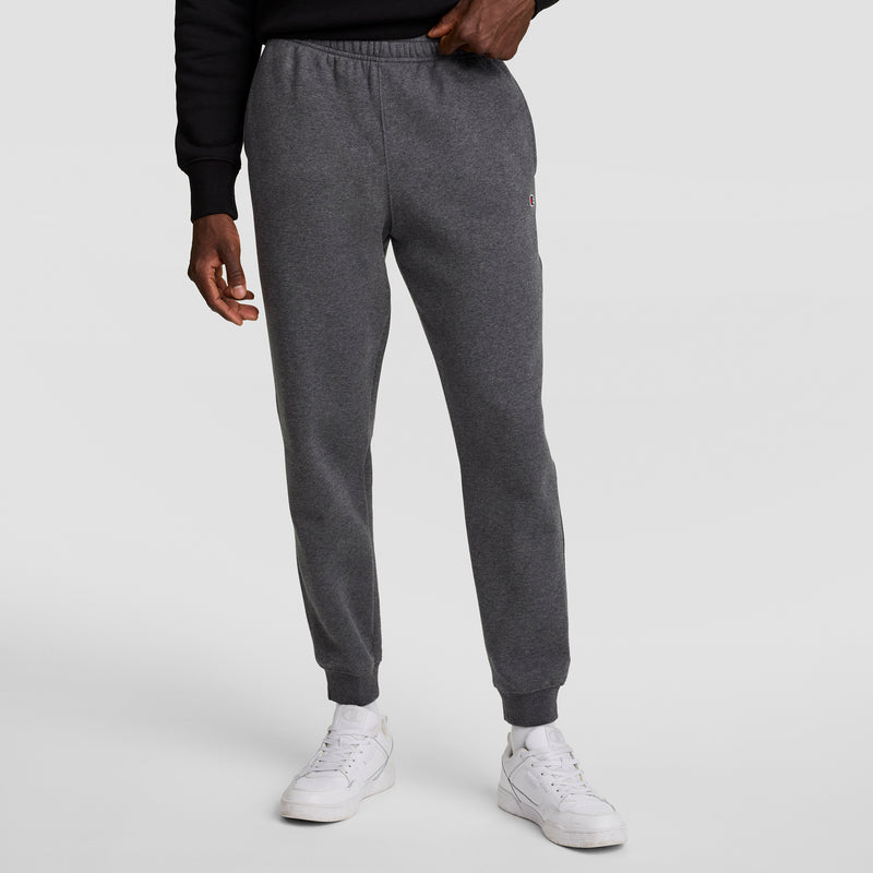 A person wears Champion Powerblend Joggers, C Logo, with a black sweatshirt and white sneakers against a plain light gray background. Only the lower half of the body is visible.