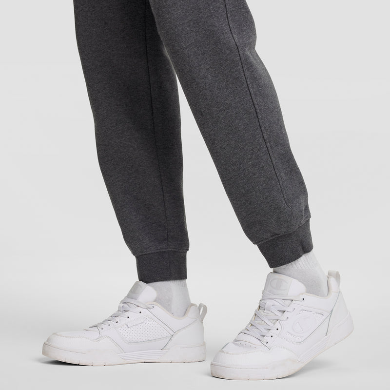 A person wearing Champion Powerblend Joggers, C Logo in dark gray, paired with white socks and white athletic sneakers, stands on a plain light background.