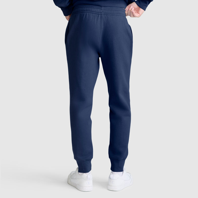 A person stands with their back to the camera, wearing Champion Powerblend Joggers with the C Logo in navy blue and white sneakers against a plain light gray background.
