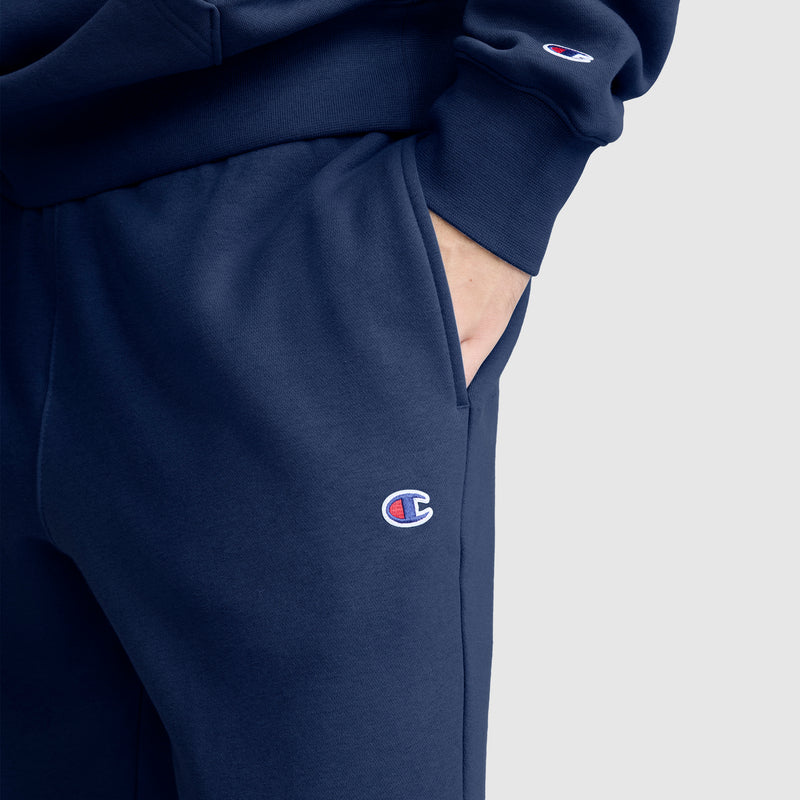A person wearing a navy blue pair of Champion Powerblend Joggers, C Logo, with their hand in the pocket. The Champion C logo is visible on both the sweatshirt sleeve and the jogger pants, showcasing the classic style of men’s joggers crafted from cozy fleece material.