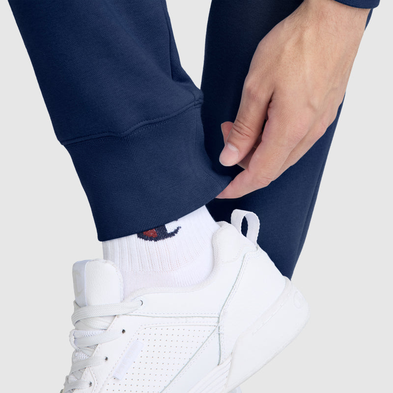 A person wearing Champion Powerblend Joggers featuring the C logo, paired with white logo socks and white athletic sneakers, adjusts the cuff of their pants.