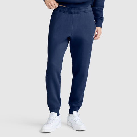 A person wearing Champion Powerblend Joggers, C Logo in navy blue cotton-blend with white sneakers stands with one hand in their pocket against a plain light gray background. The upper body is partially visible.