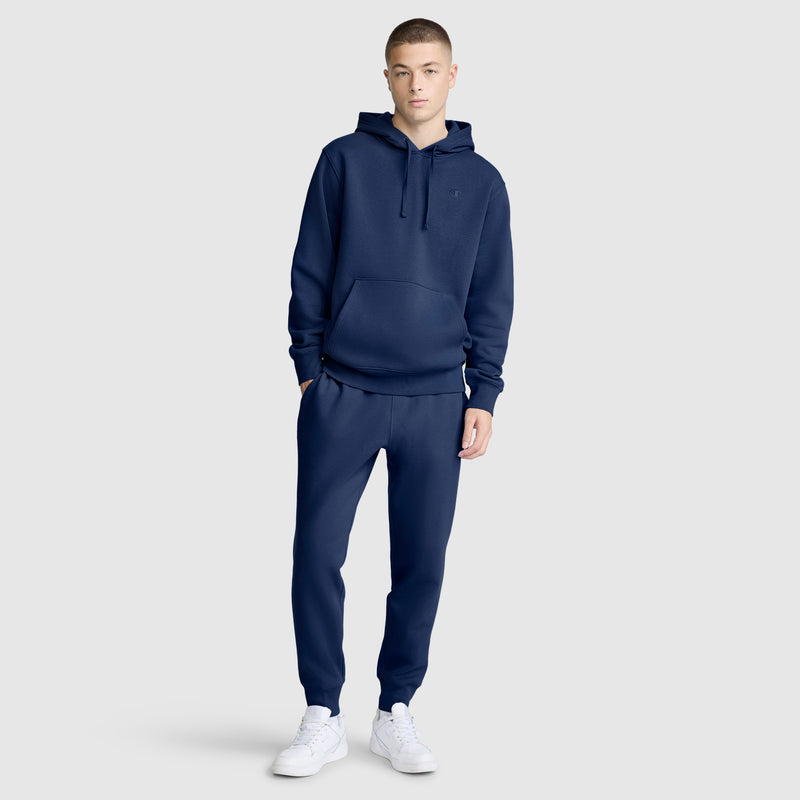 A young man wearing Champion Powerblend Joggers with the C Logo, paired with a navy blue hoodie and white sneakers, stands against a plain light gray background.