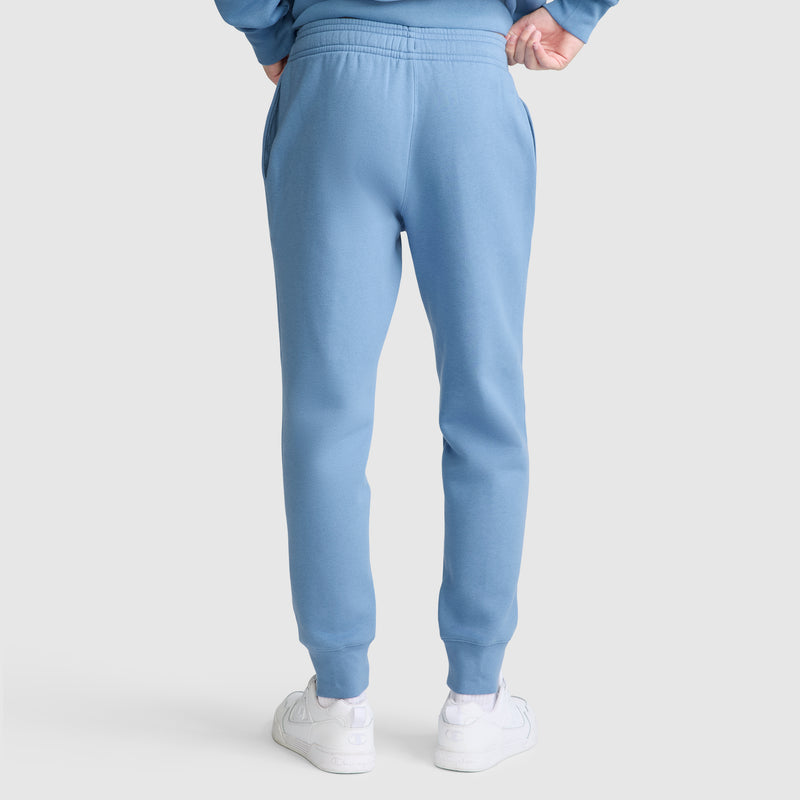 A person is shown from the back wearing Champion Powerblend Joggers, C Logo in light blue and white sneakers, standing against a plain light gray background.