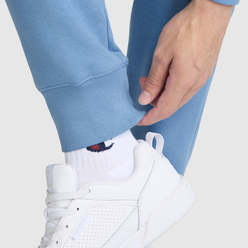Wearing Champion's Powerblend Joggers with the signature C logo, a person in light blue women's sweatpants adjusts the cuff by their ankle, their white sneakers and white socks highlighting both the comfort and iconic style of these Champion joggers.
