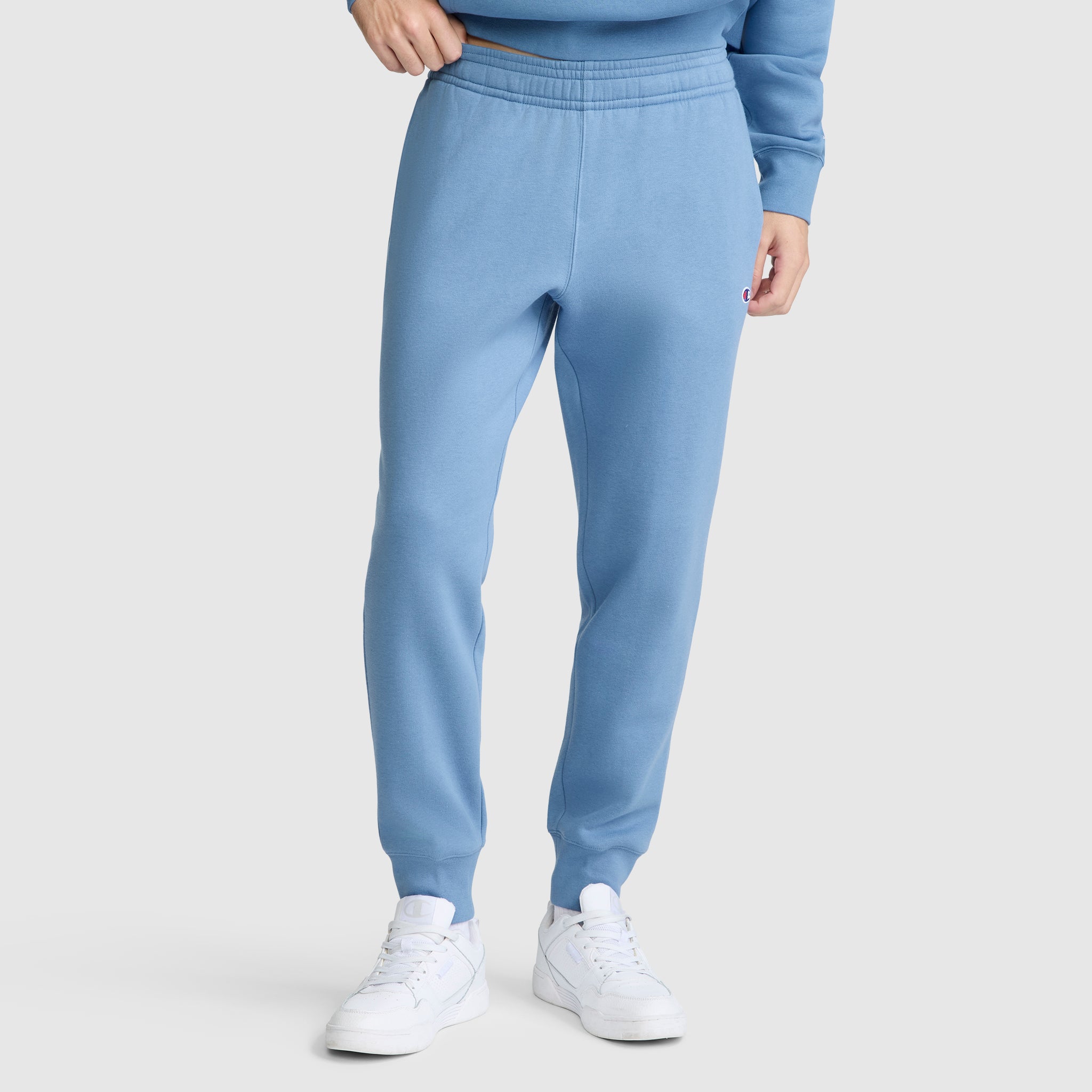 A person is shown from the waist down wearing Champion Powerblend Joggers with the C Logo in light blue, paired with a matching sweatshirt and white sneakers, standing against a plain background.
