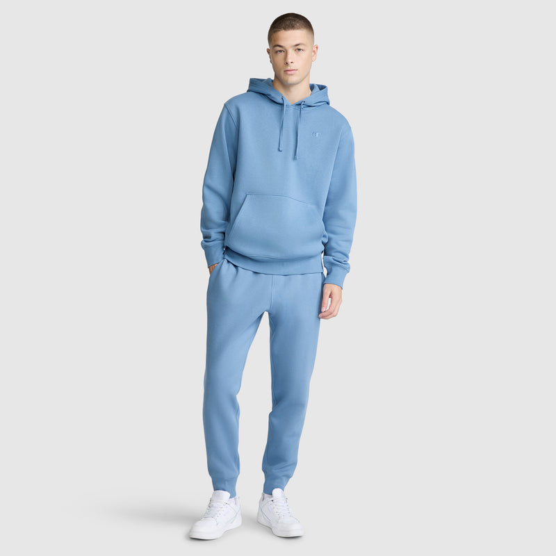 A young person wearing a matching light blue hoodie and Champion Powerblend Joggers with C Logo, paired with white sneakers, stands against a plain light gray background.